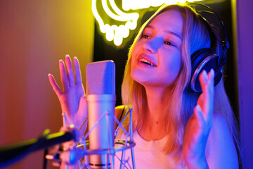 Young woman records a song into a studio microphone under neon LED lighting, blue and purple, in a...