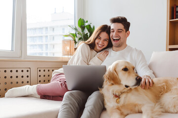 Smiling handsome boyfriend and girlfriend sitting on sofa, hugging watching video laughing