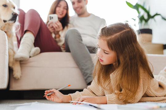 Portrait of positive attractive family, dog lying on sofa, little daughter sitting on floor drawing