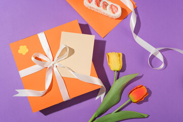Two tulip branches placed next to an orange gift box, a cake and ribbon on a purple background. Birthday concept. Copy space for advertising with a top-down perspective.