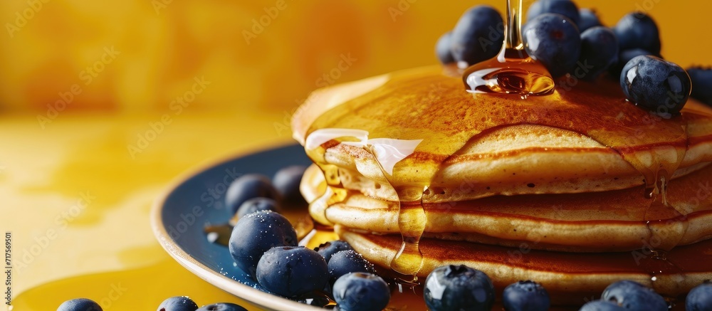 Poster Horizontal image of maple syrup pouring onto pancakes and blueberries on yellow with copy space Food organic ingredients and healthy eating. Copy space image. Place for adding text or design