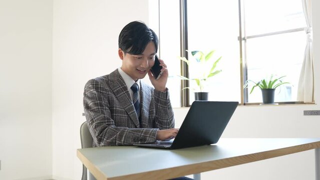 携帯で話しながらパソコンを見る若い男性