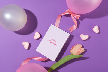 A card with the words happy birthday placed in the center of a purple background, surrounded by decorations with balloons, tulips, pink ribbons and heart-shaped marshmallows.