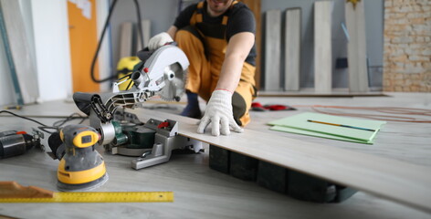 Repairman sawing laminate panels floor in room. Laying laminate around radiator pipes, installation baseboards. Skill in field repair finishing work. Trim panels with an electric jigsaw - obrazy, fototapety, plakaty