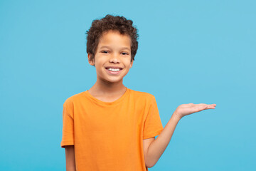 Happy boy presenting something with hand in orange shirt, blue background