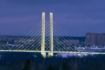 Fototapeta premium Night view of the silhouette of the illuminated Arkhangelsk bridge in Cherepovets against the background of lights of residential high-rise buildings.