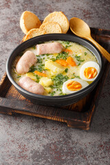 Authentic zurek a hearty Polish sour rye soup with rich white sausage and fresh veggies close-up in a bowl on the table. Vertical