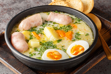 Polish zurek a rich soup soured with fermented rye starter, served with a boiled halved egg and a meaty white sausage close-up in a bowl on the table. Horizontal