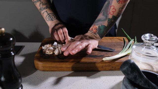 The cook is slicing garlic on a wooden chopping board in the kitchen. Mincing garlic on a wooden board with a sharp knife