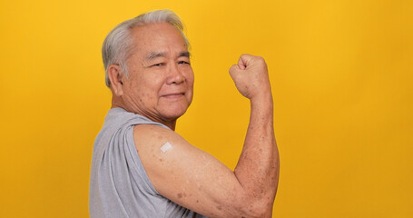Asian old man showing adhesive plaster bandage on shoulder after vaccination. Isolated on yellow...