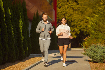 Young married couple having an outdoor fitness cardio workout. Happy husband and wife jogging on a sunny morning or evening. Smiling man and woman in sports clothes running on a green street together - obrazy, fototapety, plakaty