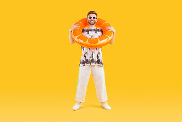 Happy handsome young man in casual summer clothes and sunglasses wearing orange inflatable beach ring on his neck and standing isolated on yellow color background. Holiday, vacation, fashion concept