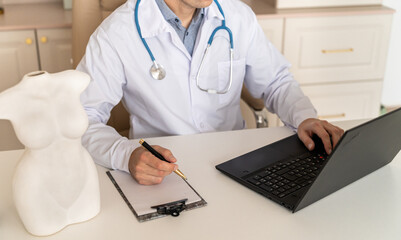 Male general practitioner taking notes in medical history of patient, writing down symptoms, complaints and anamnesis. Handsome doctor in white uniform with stethoscope working on laptop in the office - obrazy, fototapety, plakaty