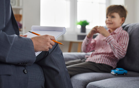 Woman psychologist or therapist with pencil and paper notepad having conversation therapy session with child kid boy who is sitting on sofa with toy car in background. Clipboard, woman's hand close up