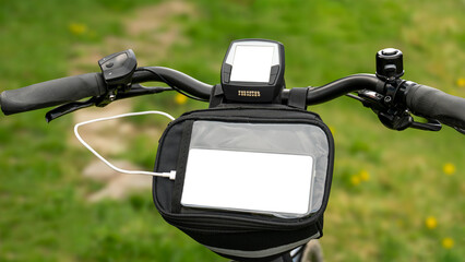 E bike handlebars with display and handlebar bag against a green blurred background. A cable comes out of the handlebar bag and leads to the smartphone. Copy space for display and cell phone.