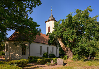 Fototapeta na wymiar Im Schatten alter Bäume steht die denkmalgeschützte Dorfkirche Wustermark am 66-Seen-Wanderweg - Inschriften wurden retuschiert
