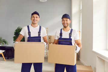 Portrait of a two young male happy smiling employee of moving service in overall standing in the living room of new house holding cardboard boxes in hands and looking cheerful at camera.