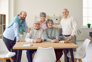 Portrait of a group of happy smiling senior people men and women sitting in nursing home at the table with books and looking cheerful at camera. Pensioners spending leisure time together.