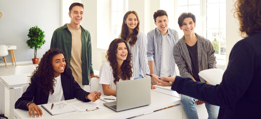 High school diverse happy smiling students at the lesson in modern classroom with laptop listening a male teacher standing back during a lesson. Education and knowledge concept. Banner.