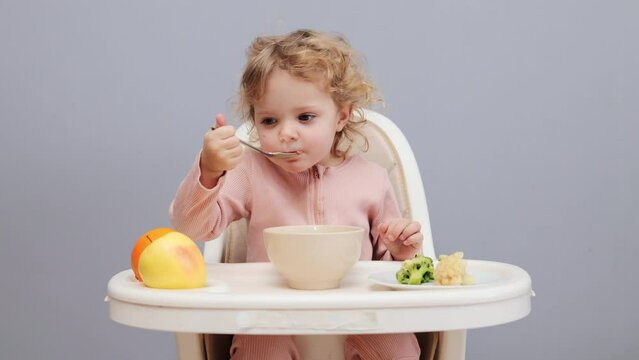 Caring for baby's food. Playtime during meals. Children's menu. Baby and dinner. Little baby girl with wavy hair eating in highchair isolated over gray background