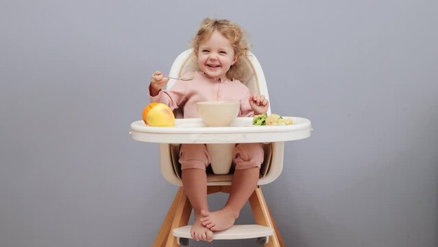 Children's health menu. Baby and diverse nutrition. Adorable little baby girl with wavy hair eating in highchair and laughing isolated over gray background