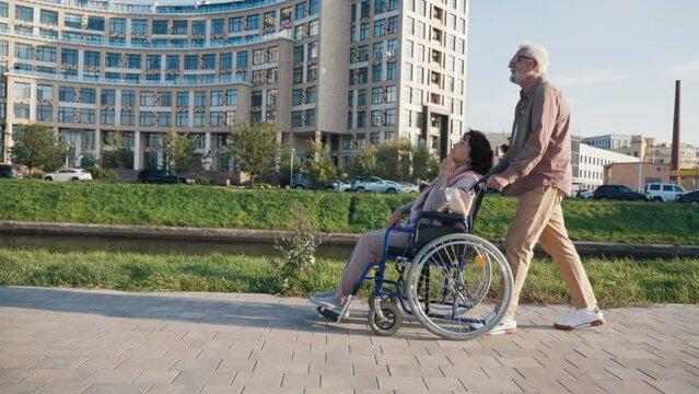 Elderly husband walking with wife in wheelchair. Senior family couple spending time in city park. Man caring woman showing love, they talking smiling. Living with physical disability, support concept.