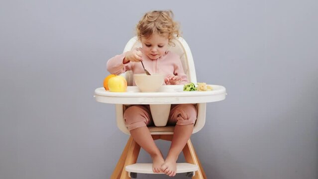 Delicious children's dishes. Healthy baby nutrition. Innocence in feeding. Hungry cute little baby girl with wavy hair eating in highchair isolated over gray background