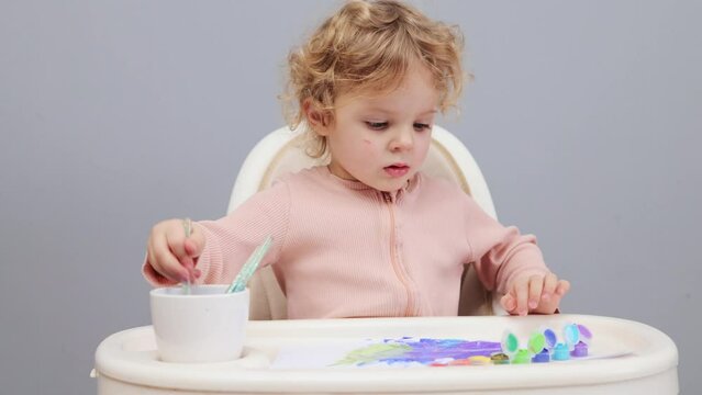 Fun with art. Small toddler artist. Kindergarten creativity. Childhood watercolor. Laughing funny curly haired blond in highchair painting with watercolor isolated over gray background