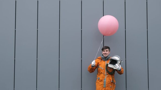 Happy astronaut wearing space suit takes space helmet off and smile while holding pink helium balloon outdoors. Surreal concept