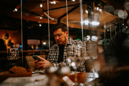 Malaga, Spain - January, 21, 2024 -  man in a plaid shirt looking intently at his smartphone in a restaurant setting with blurred lights...