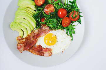 tasty breakfast - fried egg and bacon served with arugula tomato salad and fresh avocado on white plate