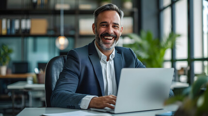 Happy, smiling, middle-aged professional business man, CEOmanager of a company, dressed in a suit, working on a laptop at his desk and laughing at work. Real unposed picture