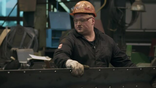 Metallurgy production worker wearing protective glasses and an orange hat. Metallurgy production worker uses grinding tool. Profession metallurgy production worker scraping off excess steel material.