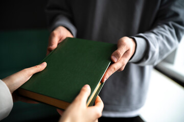 A close-up view captures the moment one person hands over a green hardcover book to another, symbolizing knowledge sharing or giving.