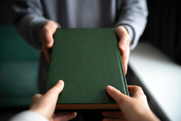 A close-up view captures the moment one person hands over a green hardcover book to another,...