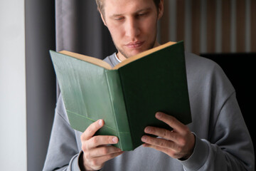 A focused young man wearing a casual grey sweater is deeply absorbed in reading a green hardcover...