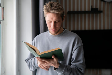 A focused young man wearing a casual grey sweater is deeply absorbed in reading a green hardcover book while seated comfortably by a window sill.