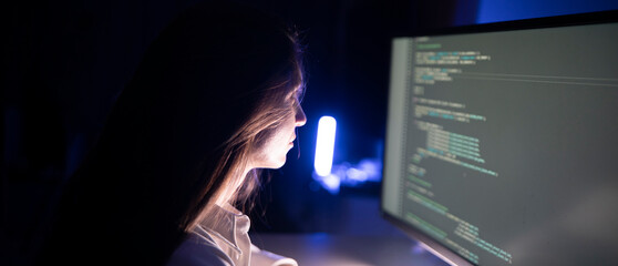 A focused woman works diligently on her coding assignment in front of a computer screen in a dimly...