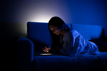 Fototapeta na wymiar A person is lying on a couch, illuminated by the soft glow of a smartphone screen as they engage with social media in the tranquility of a nighttime setting.