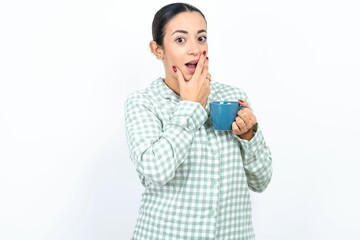 Beautiful young woman wearing green plaid pyjama and holding a cup covers mouth and looks with wonder at camera, cannot believe unexpected rumors.