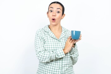 Surprised emotional Beautiful young woman wearing green plaid pyjama and holding a cup rubs palms and stares at camera with disbelief