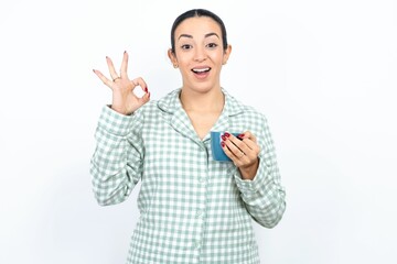 Beautiful young woman wearing green plaid pyjama and holding a cup hold hand arm okey symbol toothy...