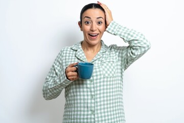 Cheerful overjoyed Beautiful young woman wearing green plaid pyjama and holding a cup reacts rising hands over head after receiving great news.