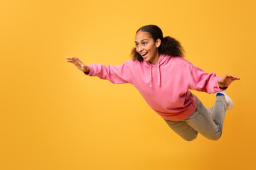 Excited black teen girl in hoodie spreading arms flying, studio