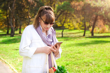 Stylish elderly caucasian brown-haired woman in sunglasses and white denim suit with linen shopping bag with vegetables, herbs and fresh baguette with mobile phone