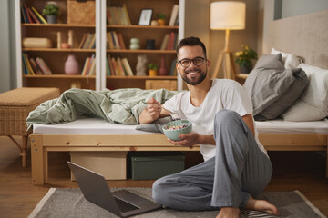 Young man using laptop while having breakfast in bedroom