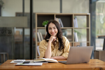 Obraz premium Attractive Asian businesswoman sitting with laptop and documents at table in office, thinking an idea, looking at camera