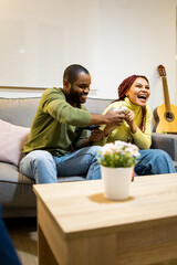 Vertical photo of a dark-skinned couple is sitting on the couch at home playing video game consoles...