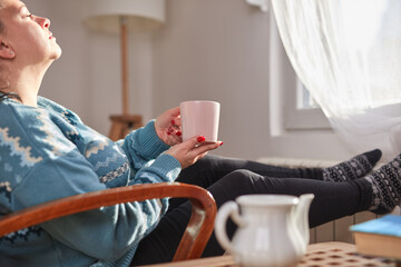 Woman drinking tea and heating on a radiator in the living room while receiving outdoor sunlight on the face.