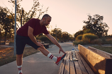 Sporty adult man stretching on a park bench after jogging in sunset time.